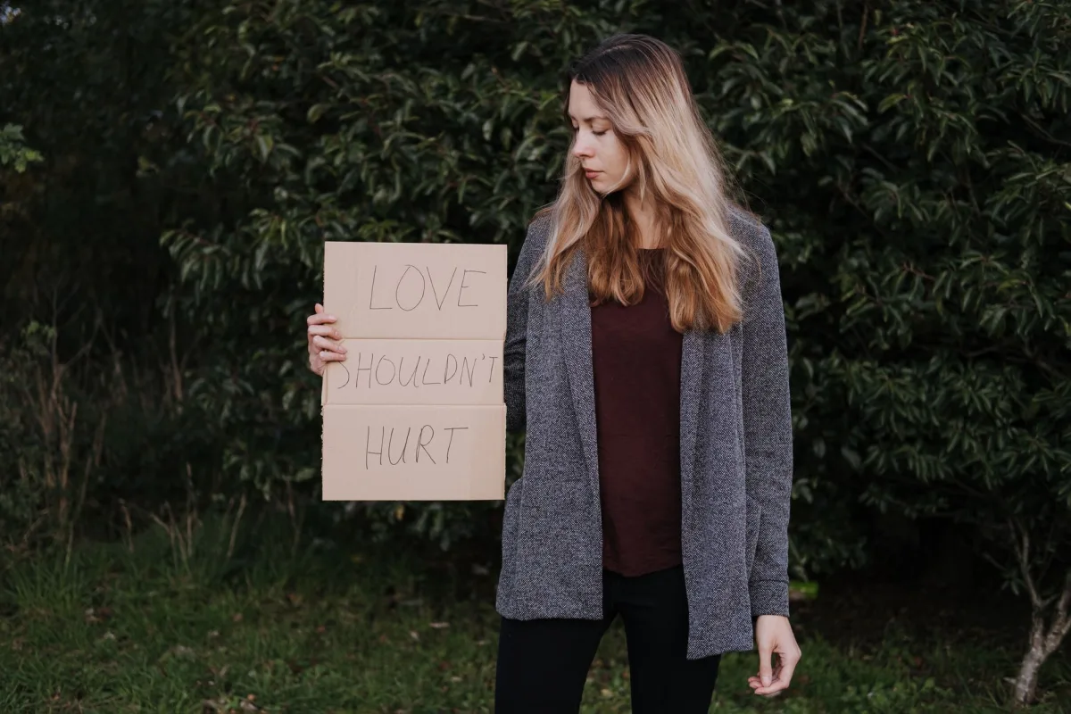 Lady in casual clothes standing near green bushes while showing poster saying Love Should Not Hurt Lady in casual clothes standing near green bushes while showing poster saying Love Should Not Hurt
